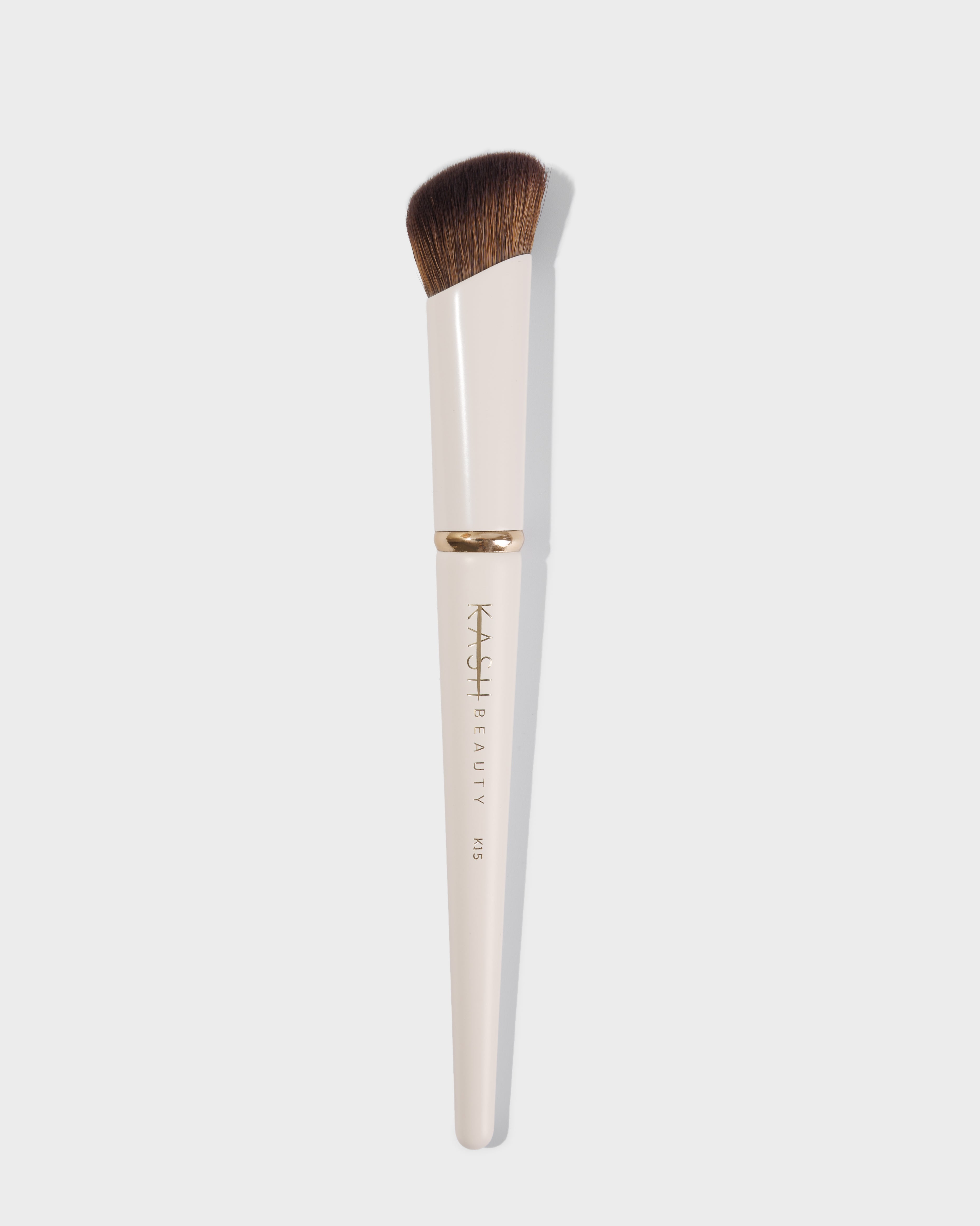 Cream-colored concealer brush with dense brown bristles by Kash Beauty, featuring gold ferrule and engraved handle, shown front view.
