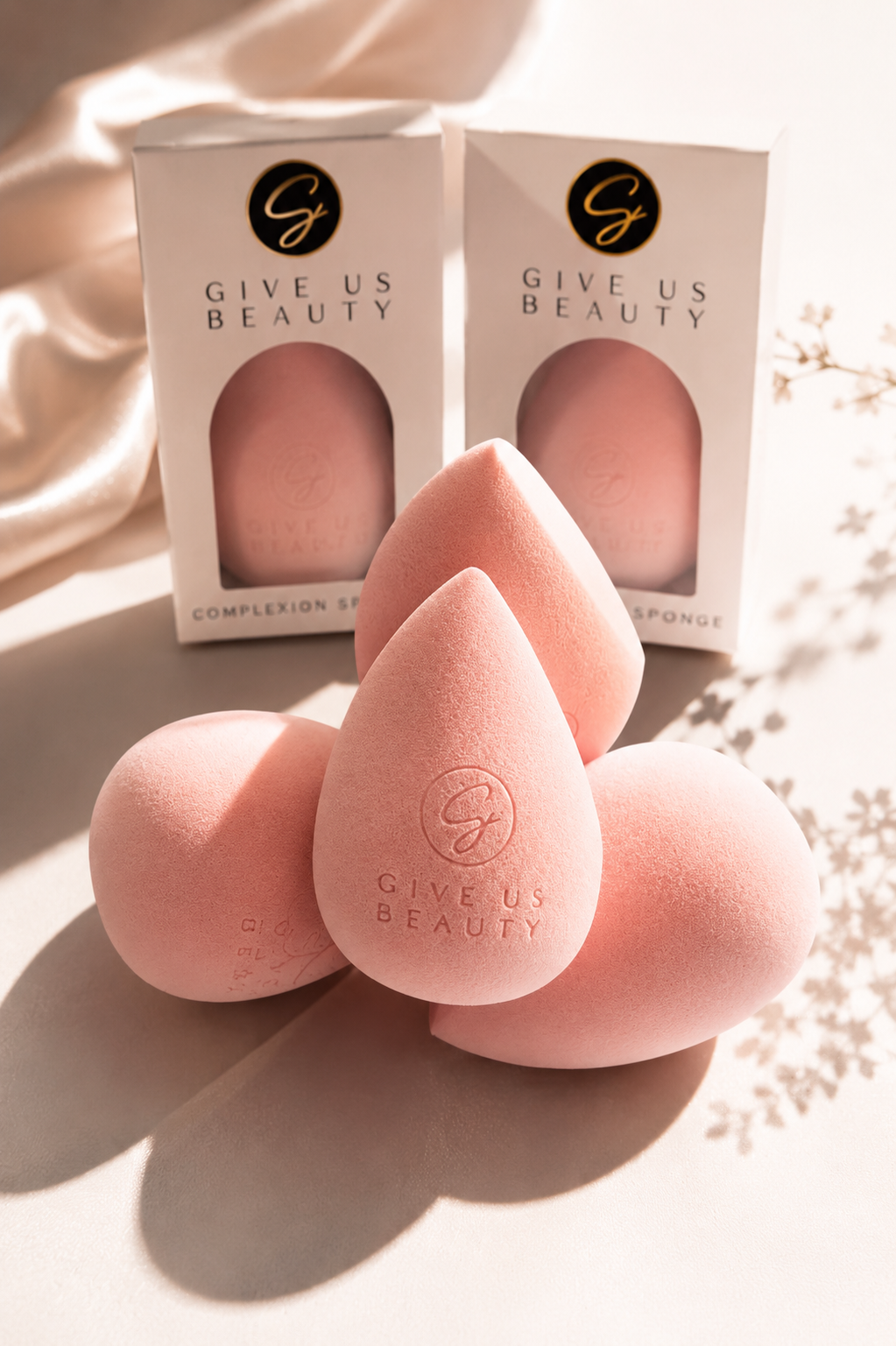 Four pink makeup blending sponges in front of two white GIVE US BEAUTY boxes, styled on a warm beige surface with soft sunlight and shadows.