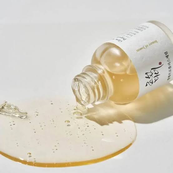 A clear glass dropper bottle tilted to pour a transparent golden liquid onto a white surface, with a small amount of the oil pooling in the foreground. The bottle has a white label with black and red text. Beauty of joseon at give us beauty