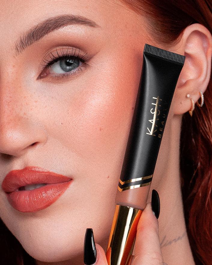 Person holding a KASHI cosmetic tube near their ear with a close-up of the product. kash beauty skin glaze honey bronze, give us beauty